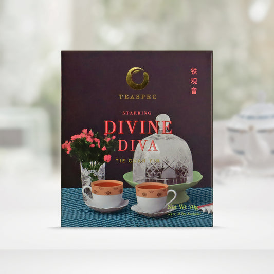 TEASPEC DIVINE DIVA Tie Guan Yin Chinese Loose Leaf Oolong Tea box with a net weight of 70g, comes in 7g Sachets x 10 per box. The front of the box design is a dark grey color with a square patterned teal tablecloth that has a textured short glass vase with small red flowers, A green cake-stand with a large glass cover and two orange and white porcelain decorative teacups next to it. The Chinese name of the tea is 铁观音