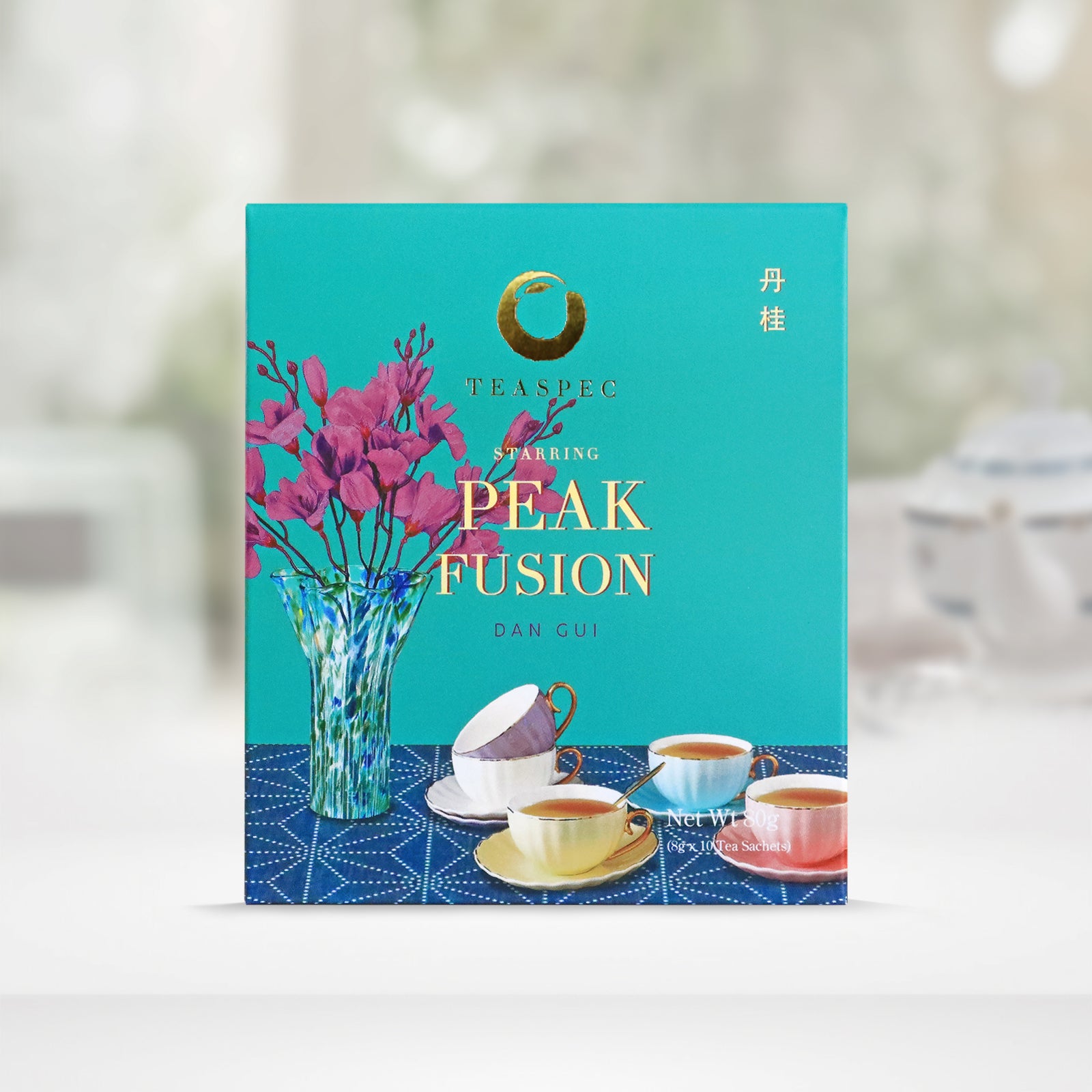 TEASPEC PEAK FUSION Dan Gui Chinese Loose Leaf Oolong Tea box with a net weight of 80g, comes in 8g Sachets x 10 per box. The front of the box design is a topaz color with a light navy tablecloth that has a glass vase with pink flowers and some white, baby blue, yellow and pink colored porcelain teacups with gold handles on it. The Chinese name of the tea is 丹桂