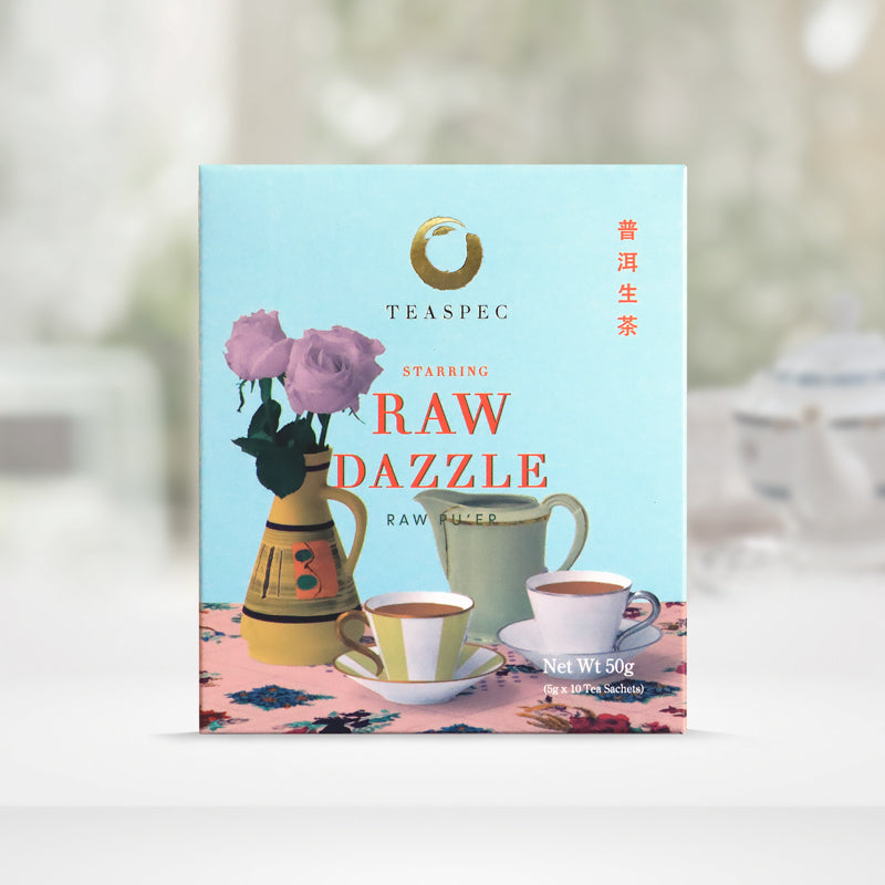 TEASPEC RAW DAZZLE Aged Raw Pu'er Chinese Loose Leaf Pu'er Tea box with a net weight of 50g, comes in 5g Sachets x 10 per box. The front of the box design is a powder blue color with a pink floral-patterned tablecloth that has a jug shaped rustic south American style vase with soft pink rose flowers. A tall light green-gray porcelain teapot with a tall handle and two white porcelain teacups in front on saucers. The Chinese name of the tea is 生普洱