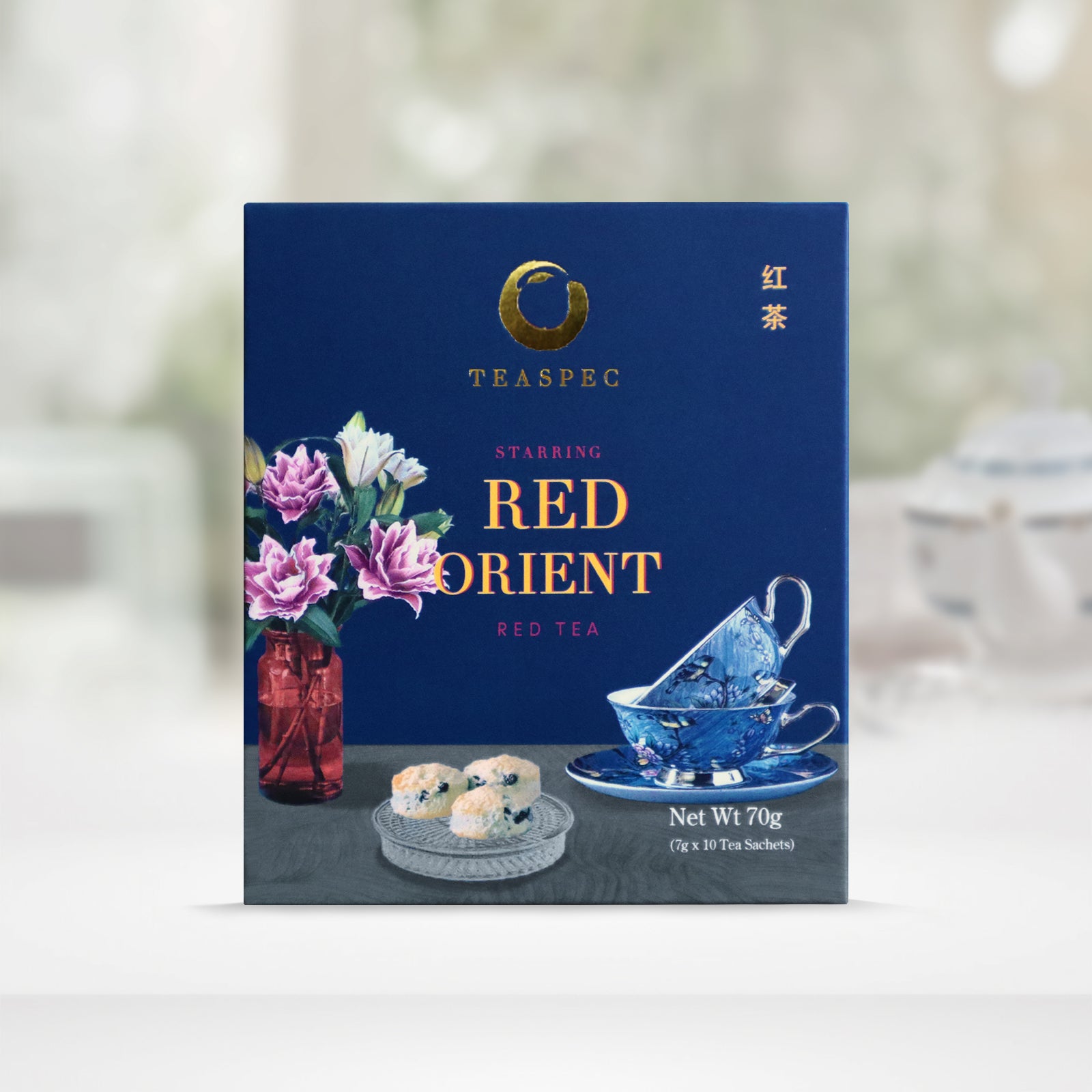 TEASPEC RED ORIENT Red Tea Chinese Loose Leaf Tea box with a net weight of 70g, comes in 7g Sachets x 10 per box. The front of the box design is a regal blue color with a gray tablecloth that has a red glass vase with pink and white rose flowers, a circular glass tray with three raisin scones and two decorative blue porcelain teacups on a saucer of the same design. The Chinese name of the tea is 红茶