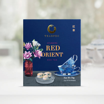 TEASPEC RED ORIENT Red Tea Chinese Loose Leaf Tea box with a net weight of 70g, comes in 7g Sachets x 10 per box. The front of the box design is a regal blue color with a gray tablecloth that has a red glass vase with pink and white rose flowers, a circular glass tray with three raisin scones and two decorative blue porcelain teacups on a saucer of the same design. The Chinese name of the tea is 红茶