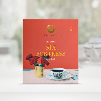 TEASPEC SIX FORTRESS Liu Bao Chinese Loose Leaf pure black Tea box with a net weight of 100g, comes in 10g Sachets x 10 per box. The front of the box design is a chestnut red color with a vanilla modern patterned tablecloth that has a yellow short porcelain vase with red flowers. Next to it is a traditional single color floral design porcelain teacup on a saucer. The Chinese name of the tea is  六堡