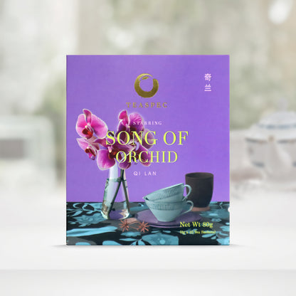 TEASPEC SONG OF ORCHID Qi Lan Chinese Loose Leaf Oolong Tea box with a net weight of 80g, comes in 8g Sachets x 10 per box. The front of the box design is a Lavander purple color with a blue-black floral pattern tablecloth with slim neck tall triangle shaped glass vase with the national flower of Singapore. Papilionanthe Miss Joaquim, an orchid. Two light blue porcelain teacups in front on saucer with a dark cup behind. The Chinese name of the tea is 奇兰