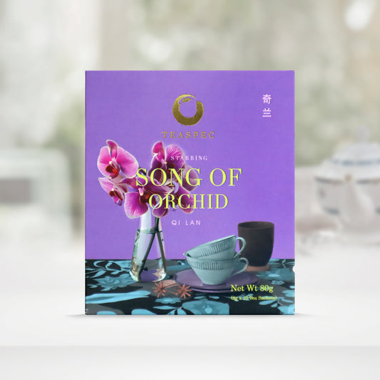 TEASPEC SONG OF ORCHID Qi Lan Chinese Loose Leaf Oolong Tea box with a net weight of 80g, comes in 8g Sachets x 10 per box. The front of the box design is a Lavander purple color with a blue-black floral pattern tablecloth with slim neck tall triangle shaped glass vase with the national flower of Singapore. Papilionanthe Miss Joaquim, an orchid. Two light blue porcelain teacups in front on saucer with a dark cup behind. The Chinese name of the tea is 奇兰