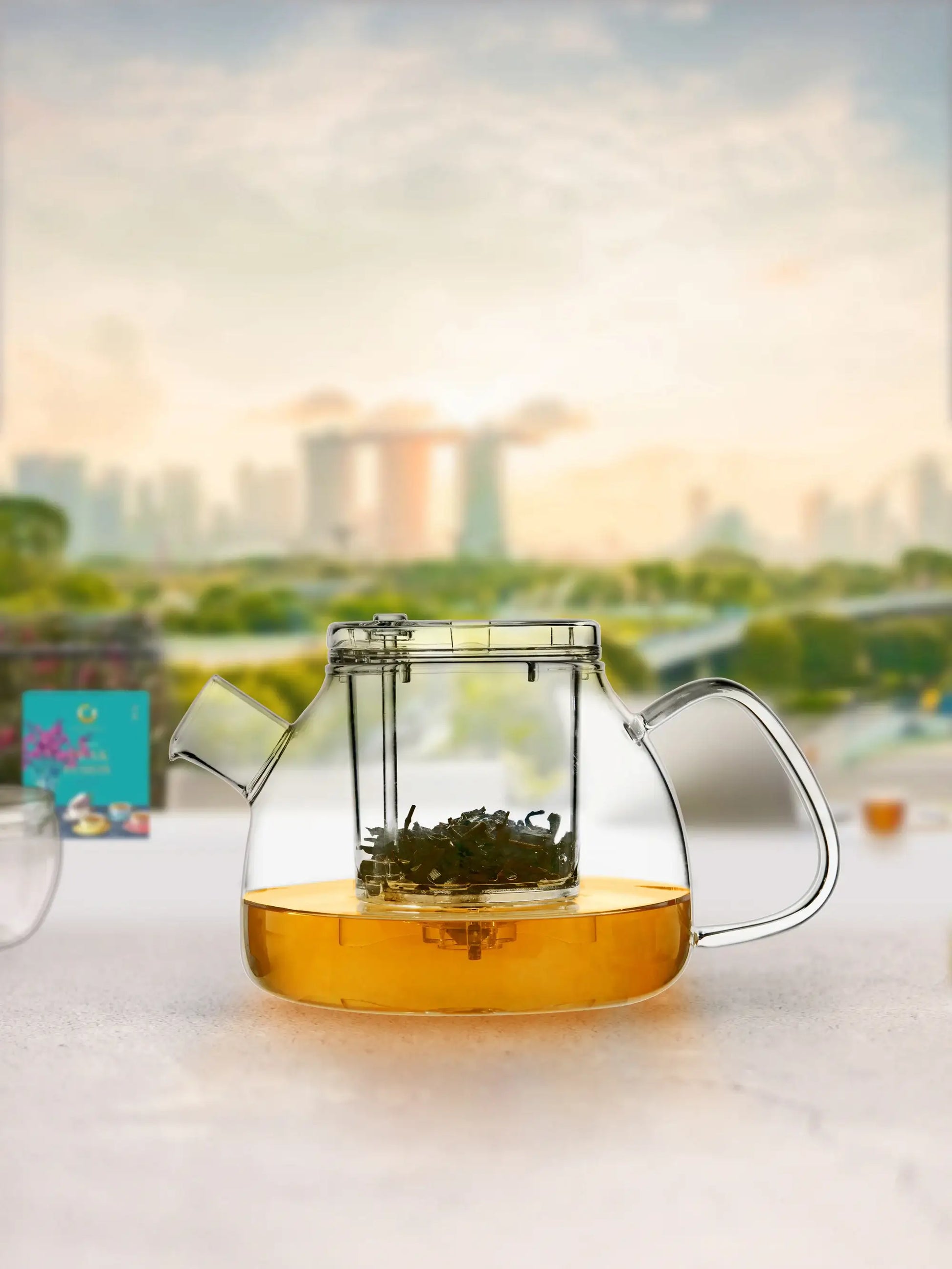 TEASPEC's Lazy Pour Glass teapot with hot tea with a Singapore marina bays sands background view. A box of PEAK FUSION also visible.