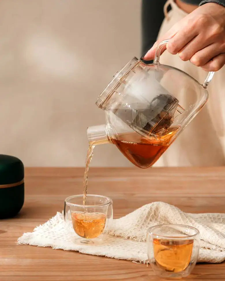 TEASPEC's Lazy Pour Glass Teapot 900ml pouring tea into a small Easy sip double walled glass teacup on a wooden countertop with a cloth below.